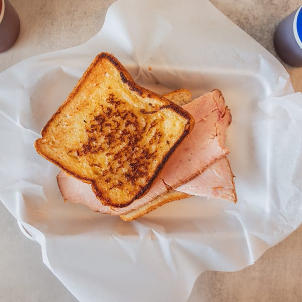 A grilled ham sandwich on toasted bread, served in a paper-lined basket with two sauce bottles.