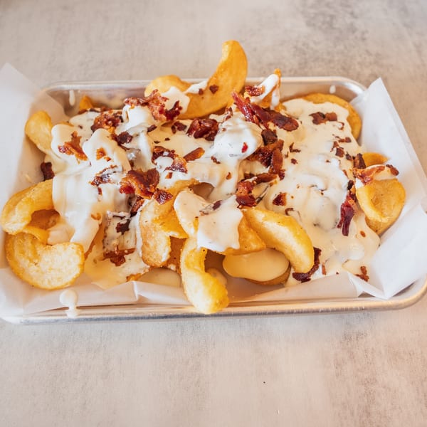 Crispy golden fries topped with creamy white sauce and crumbled bacon bits in a metal tray.
