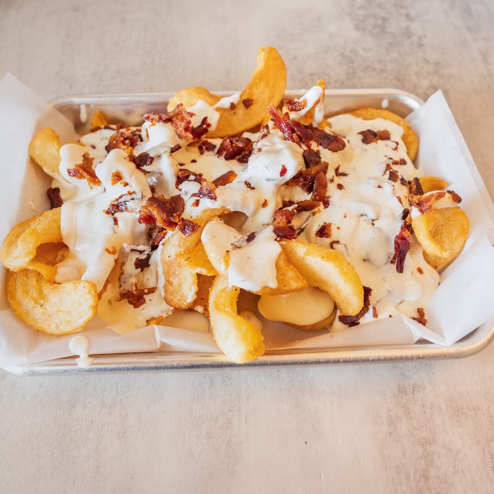 Crispy golden fries topped with creamy white sauce and crumbled bacon bits in a metal tray.