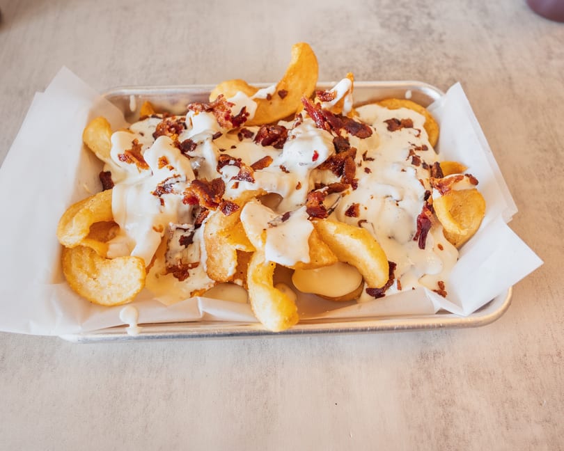 Crispy golden fries topped with creamy white sauce and crumbled bacon bits in a metal tray.