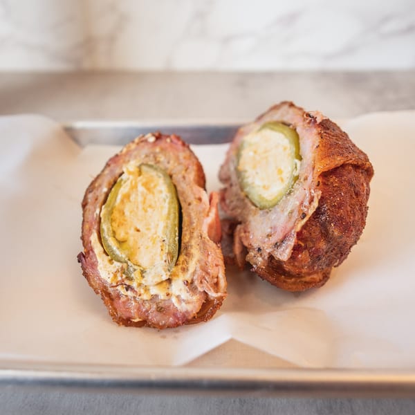 Two bacon-wrapped, cheese-stuffed jalapeño poppers, cut in half, on a white paper-lined tray.