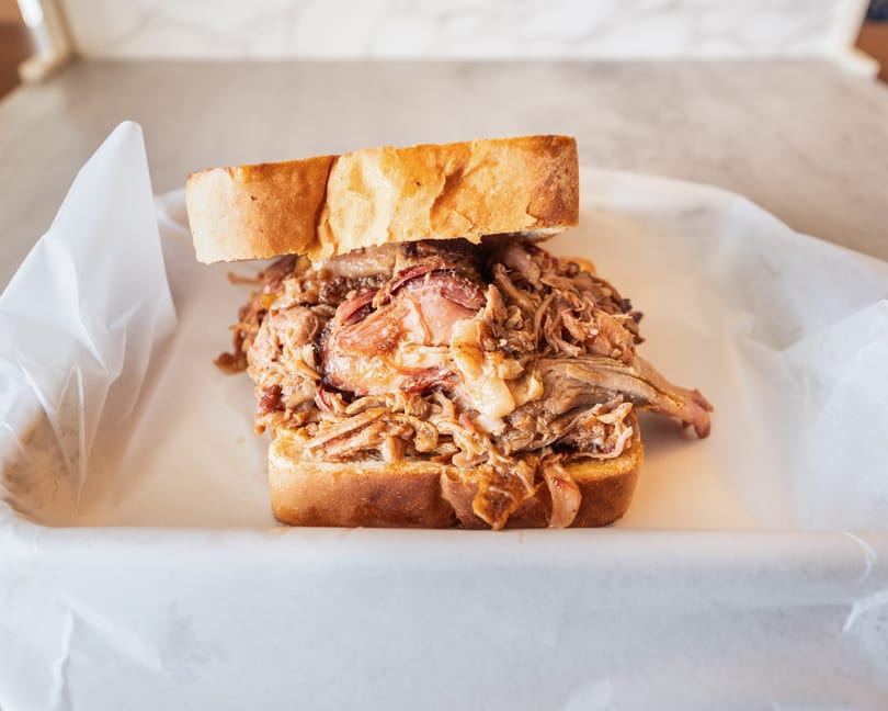 A close-up of a pulled pork sandwich on white bread, served in a paper-lined basket.
