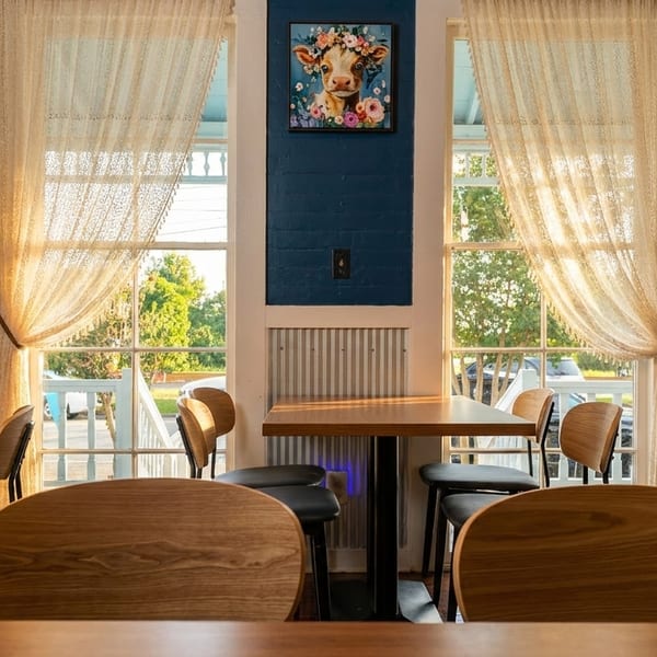 Restaurant interior with wooden tables and chairs, blue walls, and lace curtains on sunlit windows.