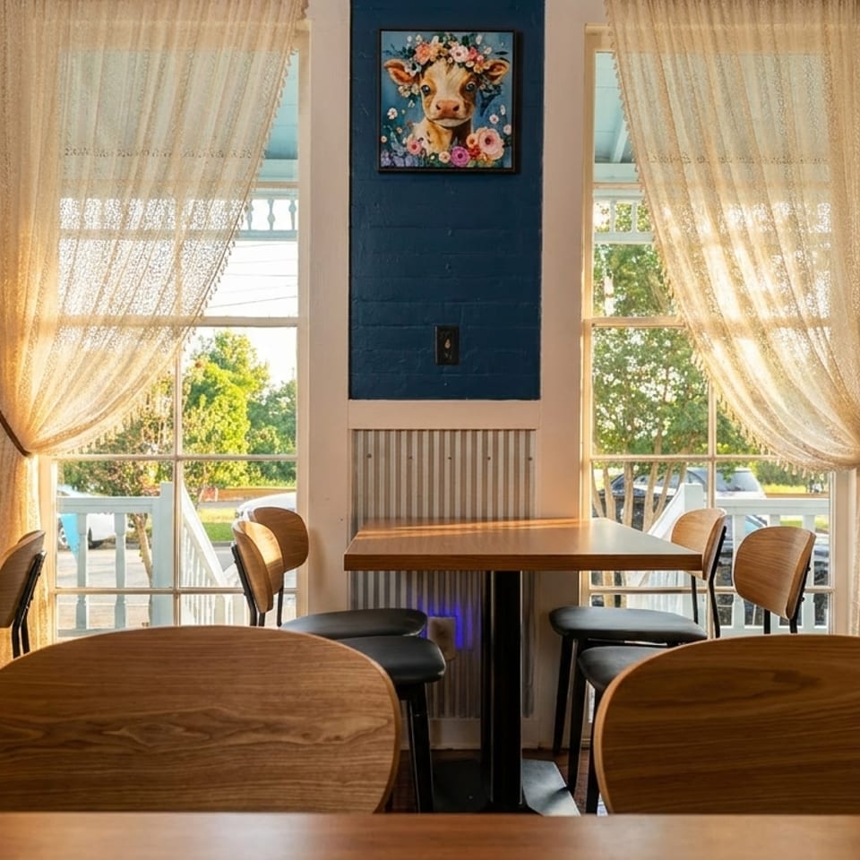 Restaurant interior with wooden tables and chairs, blue walls, and lace curtains on sunlit windows.
