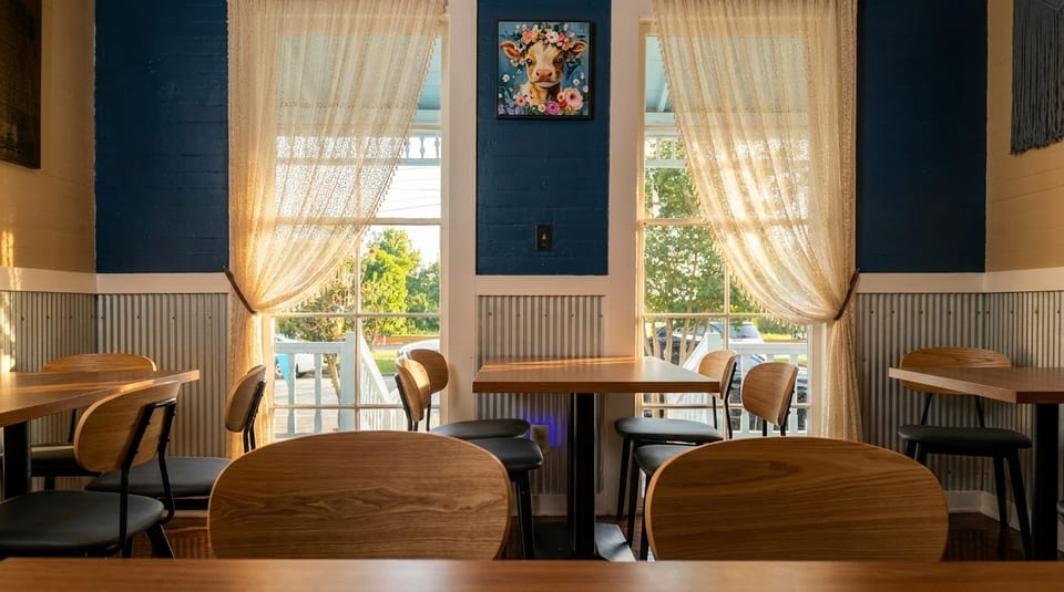Restaurant interior with wooden tables and chairs, blue walls, and lace curtains on sunlit windows.