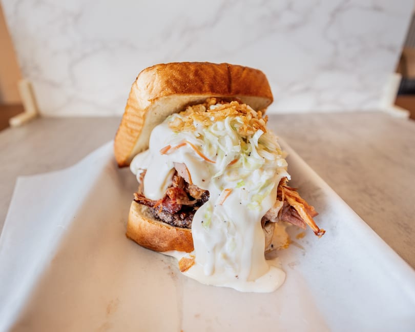 A pulled pork sandwich with coleslaw and crispy onions on Texas toast, served on parchment paper.