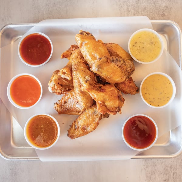 A metal tray with a pile of crispy chicken wings surrounded by six small cups of dipping sauces.
