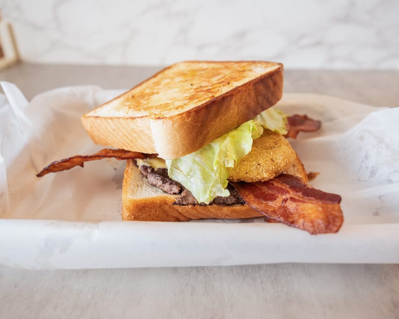 A toasted sandwich with bacon, lettuce, a fried green tomato, and a burger patty.
