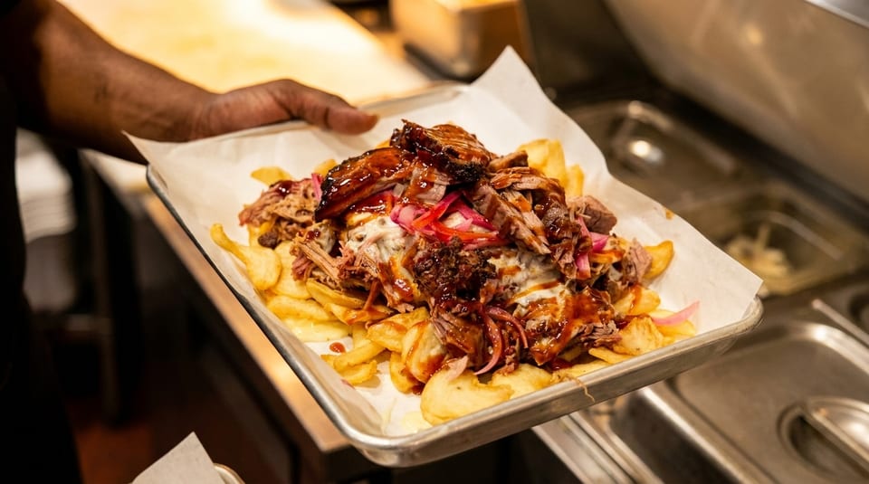 A hand holds a tray of loaded fries topped with pulled pork, BBQ sauce, and pickled onions.