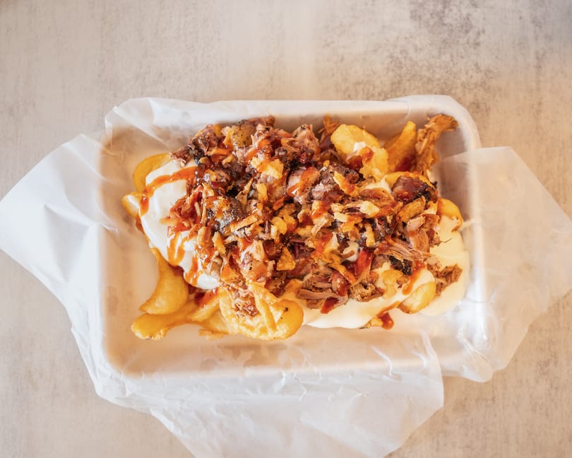 Loaded fries with pulled pork, white sauce, crispy onions, and BBQ drizzle in a white paper tray.