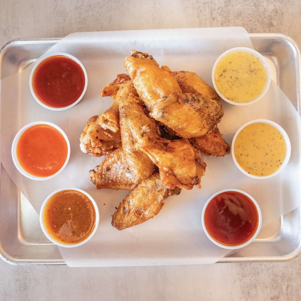 A metal tray with a pile of crispy chicken wings surrounded by six small cups of dipping sauces.