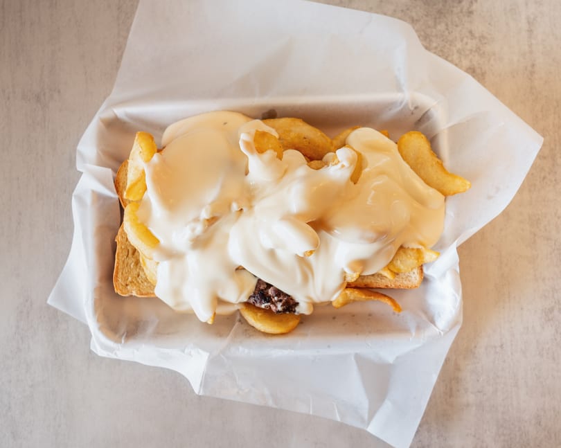 Top-down view of a burger patty and fries on toast, smothered in white cheese sauce.