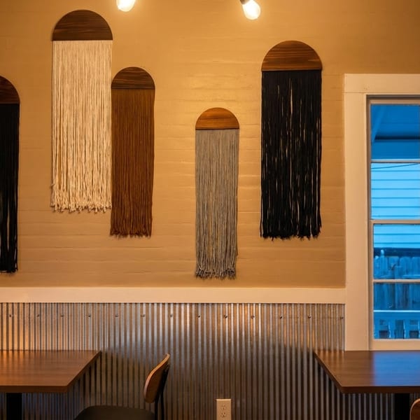 Restaurant interior with macrame wall hangings, wooden tables, chairs, and a window.