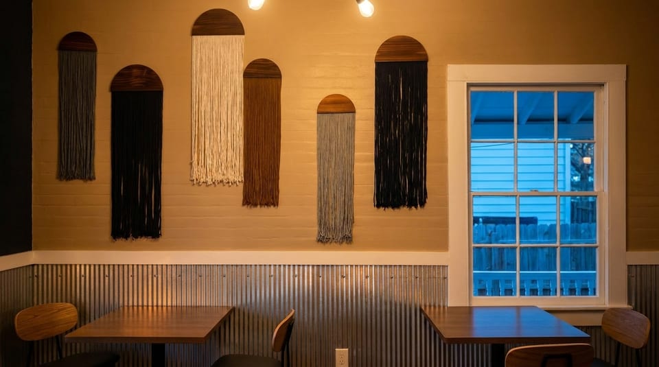 Restaurant interior with macrame wall hangings, wooden tables, chairs, and a window with blue light.